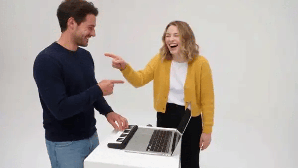 A woman in a mustard cardigan pushes her face into the lens and laughs while a man in a navy sweater keeps playing the mini keyboard behind her