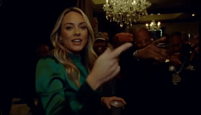 A woman in a green satin top reaches toward the camera in the chandelier room, then covers her face laughing