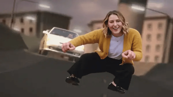 The woman in the mustard cardigan rides the invisible motorbike in mid-air, glances back at the silver car and leaps up with her legs spread