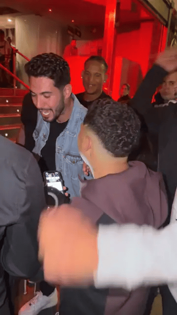 Un hombre con chaqueta vaquera levanta el brazo y se lleva una mano a la cabeza mientras la multitud lo aclama en una entrada iluminada en rojo