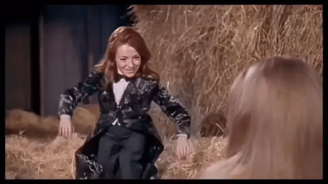 A red-haired woman in a velvet tailcoat throws her arms open and tiptoes across the hay in the barn