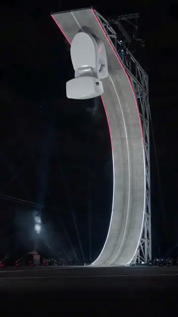 A white toilet flies off the top of the ramp upside down, flips right side up on thruster jets and lands in white smoke