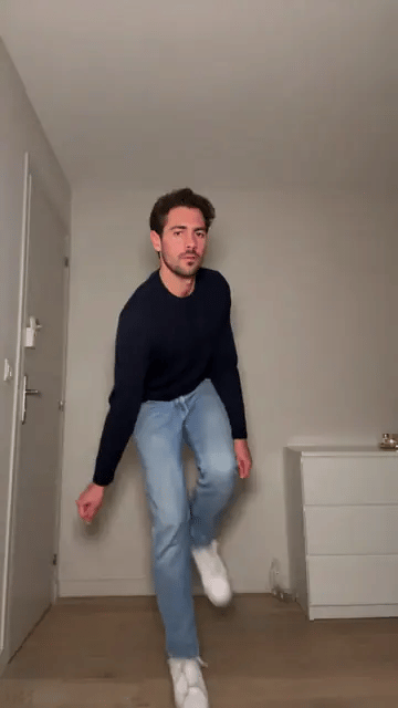 A man in a navy sweater and jeans hops on one foot, knocks on an invisible door and flings his arms wide in a bare white room