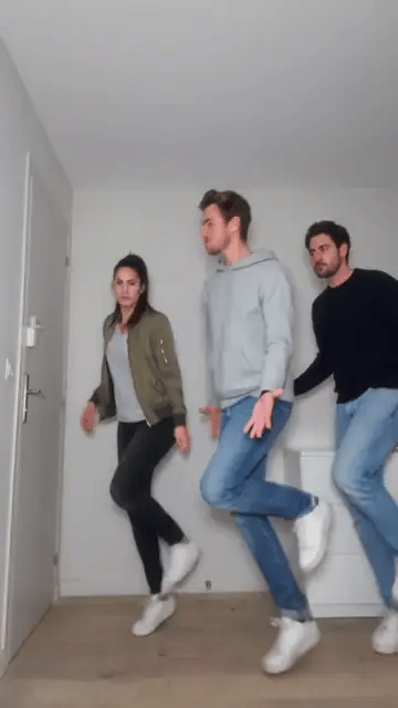 Three friends in a green jacket, a grey hoodie and a navy sweater skip sideways and kick back in sync in a bare white room